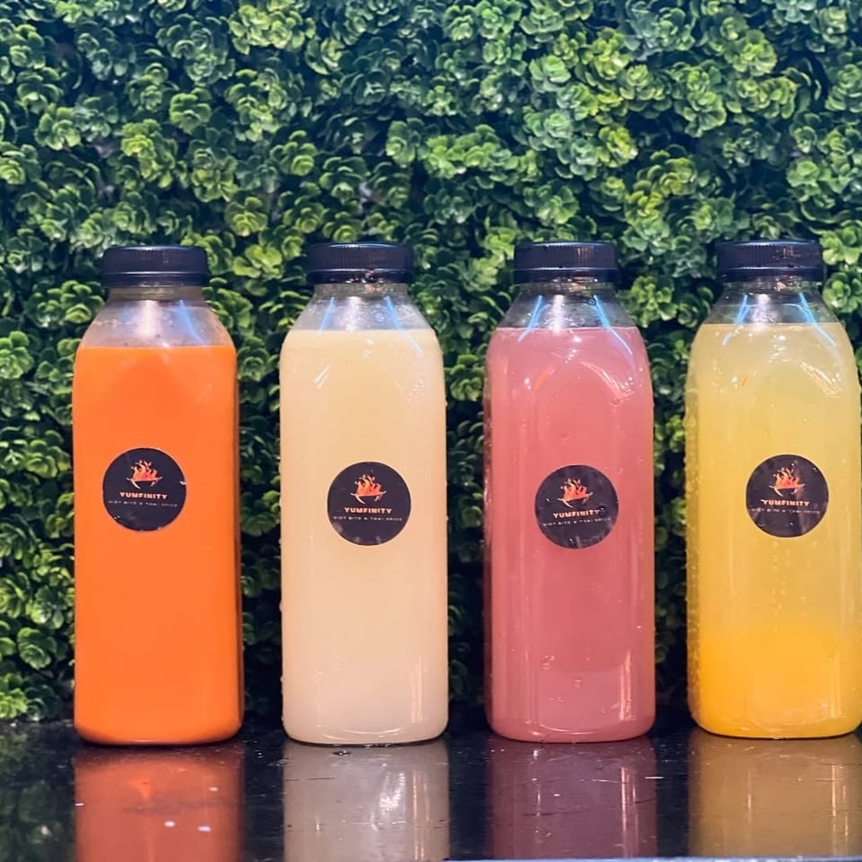 Four bottled Yumfinity drinks in orange, white, pink, and yellow against a green leafy wall.