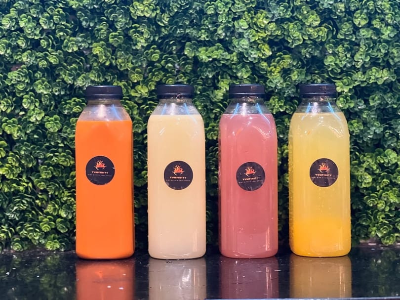Four bottled Yumfinity drinks in orange, white, pink, and yellow against a green leafy wall.