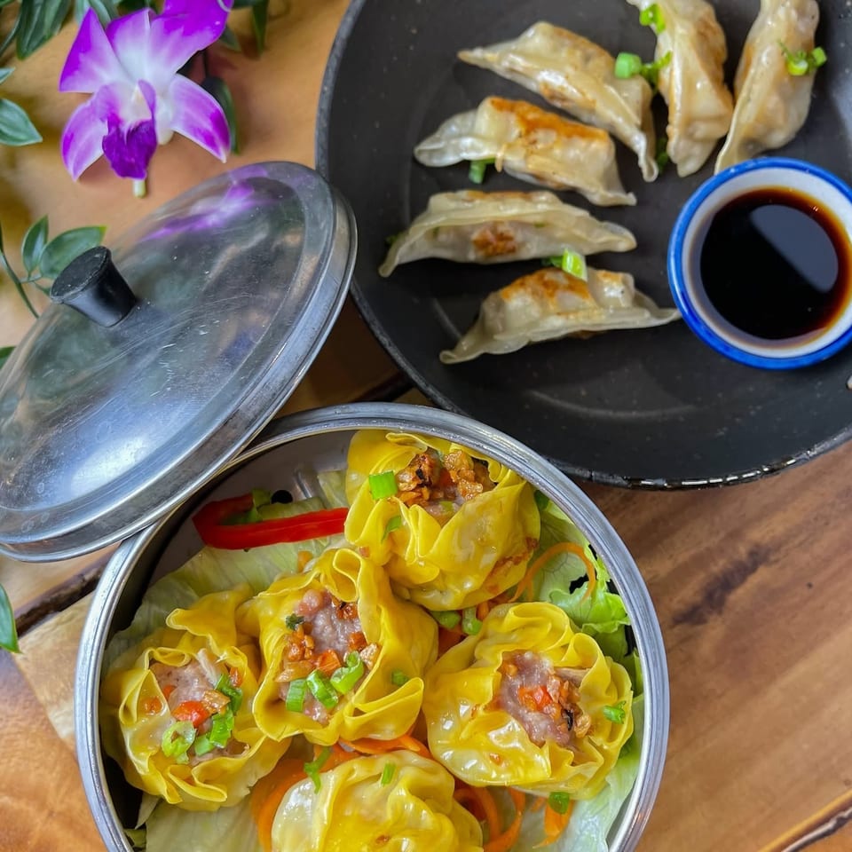 Steamed siu mai in a metal container and pan-fried dumplings with soy sauce on a wooden table.