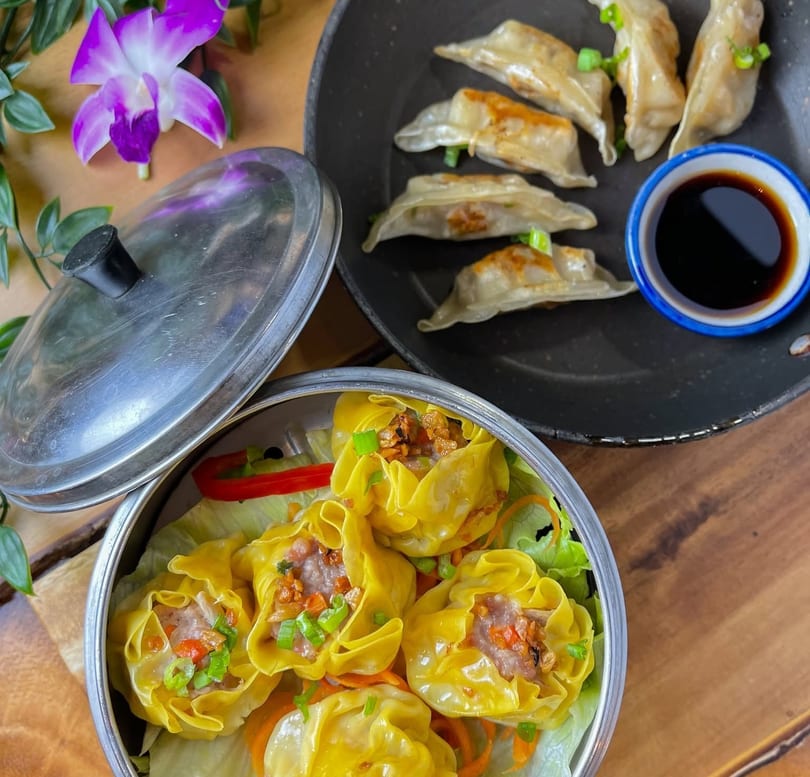 Steamed siu mai in a metal container and pan-fried dumplings with soy sauce on a wooden table.