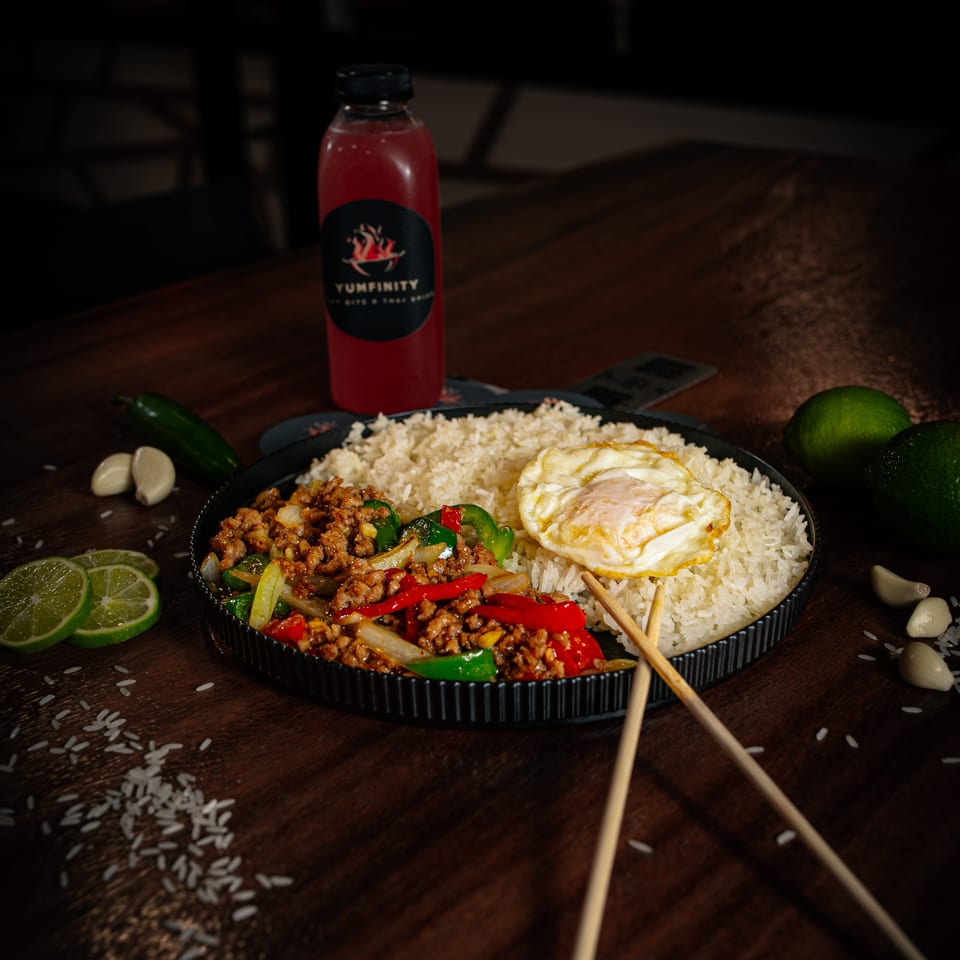 Thai basil chicken with rice and fried egg, limes, garlic, and a Yumfinity drink on a wooden table.