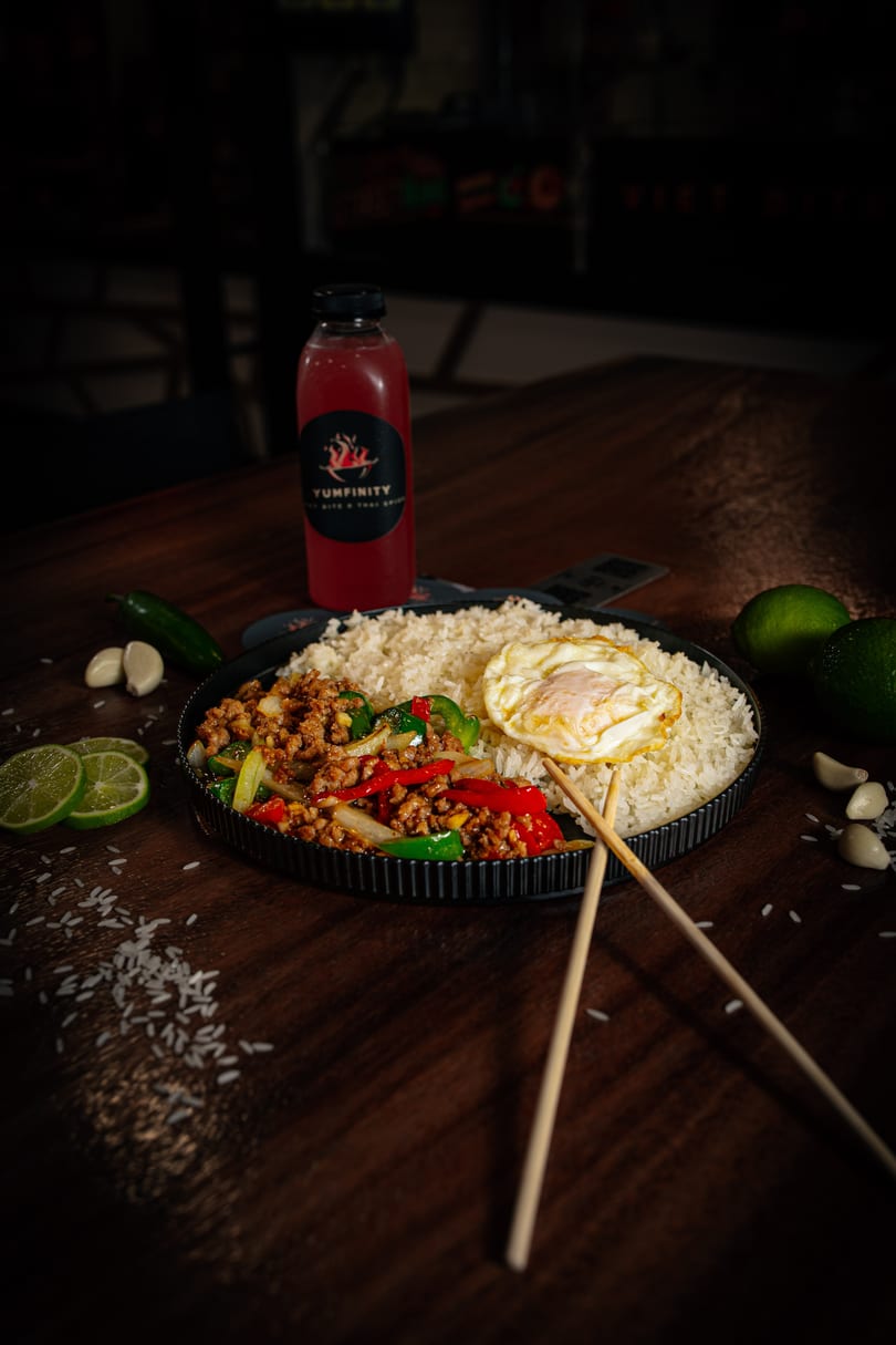 Thai basil chicken with rice and fried egg, limes, garlic, and a Yumfinity drink on a wooden table.