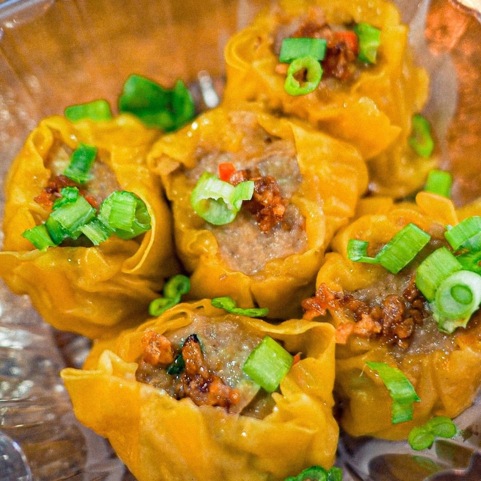 Close-up of several steamed dumplings topped with green onions and chili flakes in a plastic container.