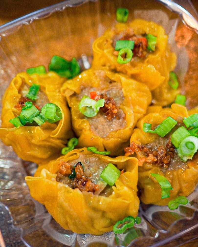 Close-up of several steamed dumplings topped with green onions and chili flakes in a plastic container.