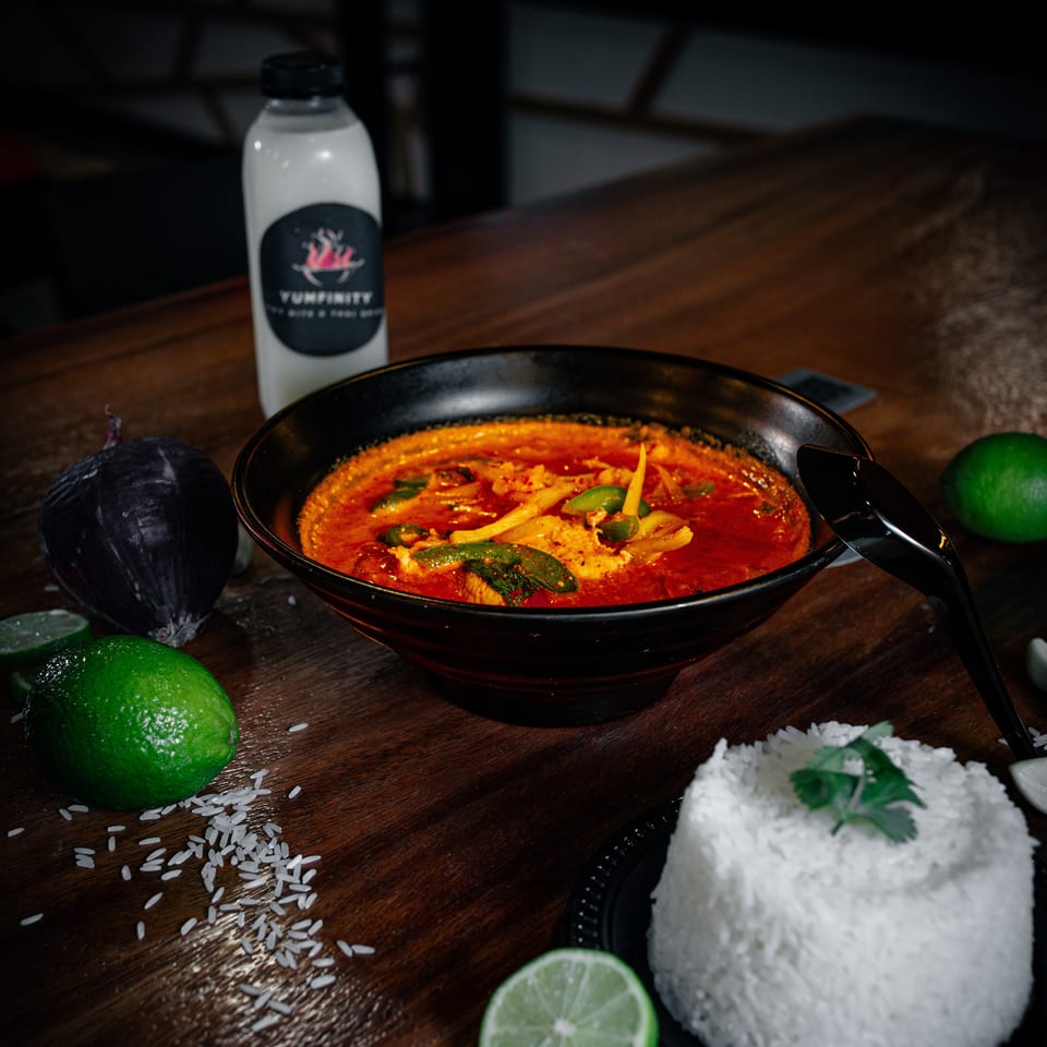 A bowl of red curry with vegetables, rice, limes, and a Yumfinity drink on a wooden table.