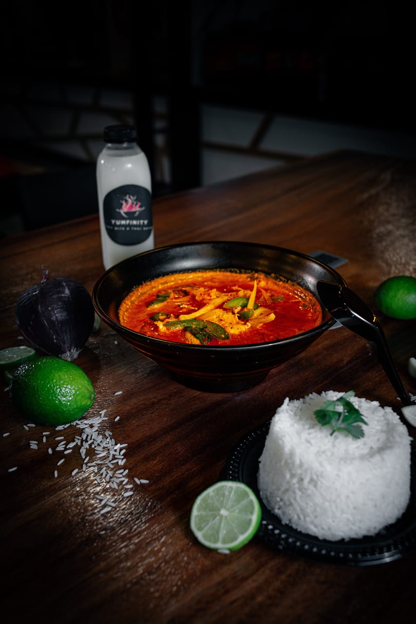 A bowl of red curry with vegetables, rice, limes, and a Yumfinity drink on a wooden table.