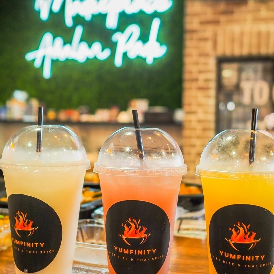 Three Yumfinity drinks, yellow, orange, and clear, on a table with a neon sign in the background.