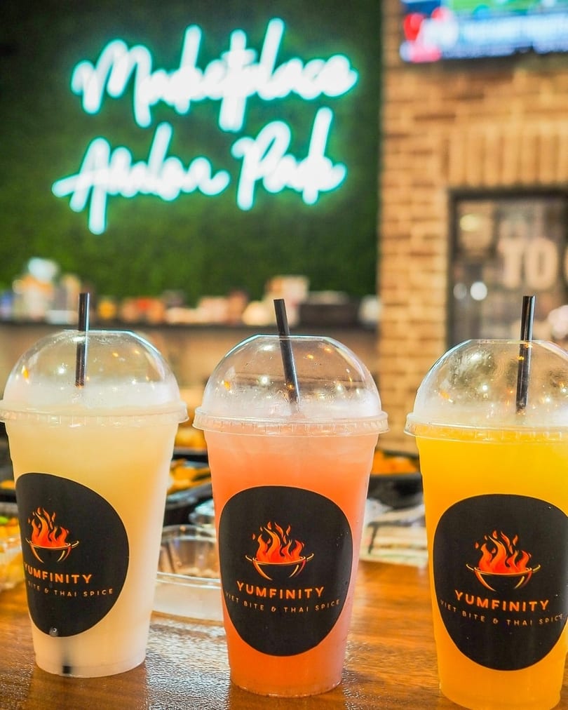 Three Yumfinity drinks, yellow, orange, and clear, on a table with a neon sign in the background.