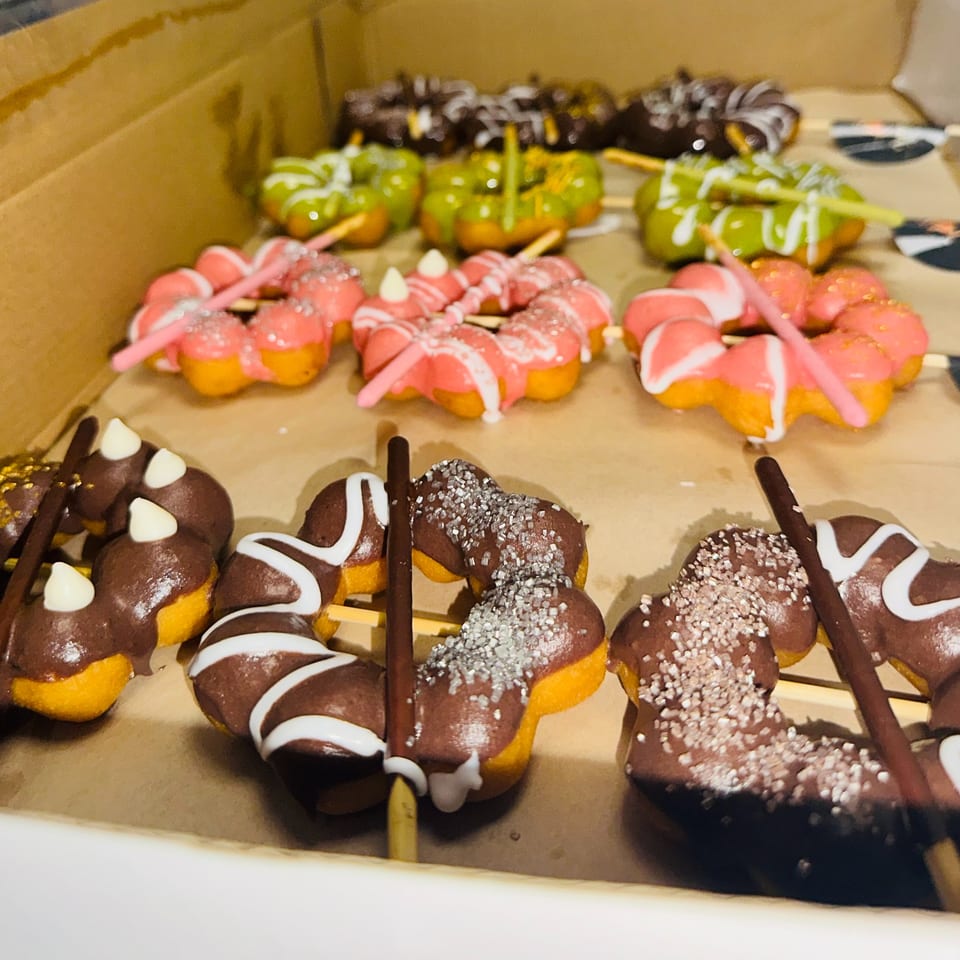 A box of colorful mochi donuts on sticks, with chocolate, pink, and green frosting.