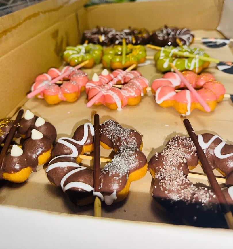 A box of colorful mochi donuts on sticks, with chocolate, pink, and green frosting.
