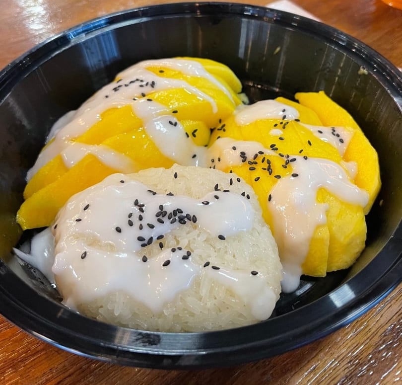 Mango sticky rice dessert with coconut milk and black sesame seeds in a black bowl.