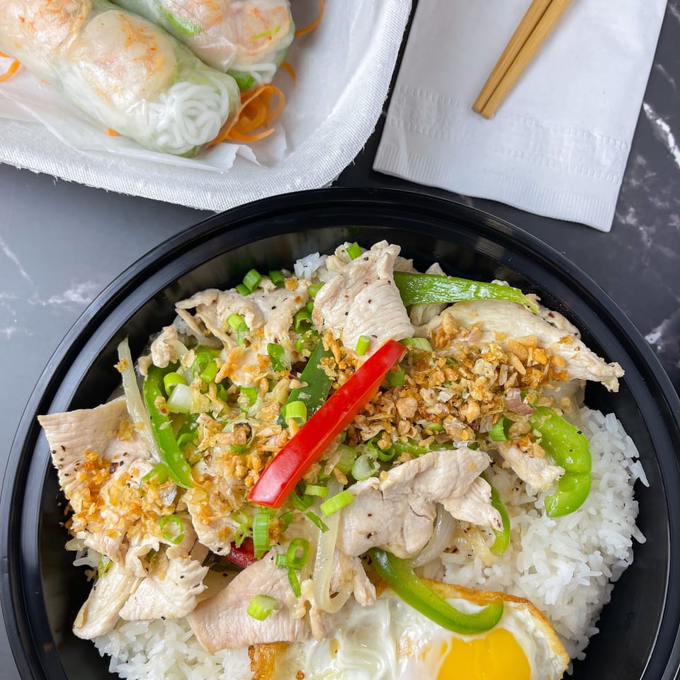 Chicken and rice bowl with a fried egg, spring rolls, and peanut sauce on a dark surface.