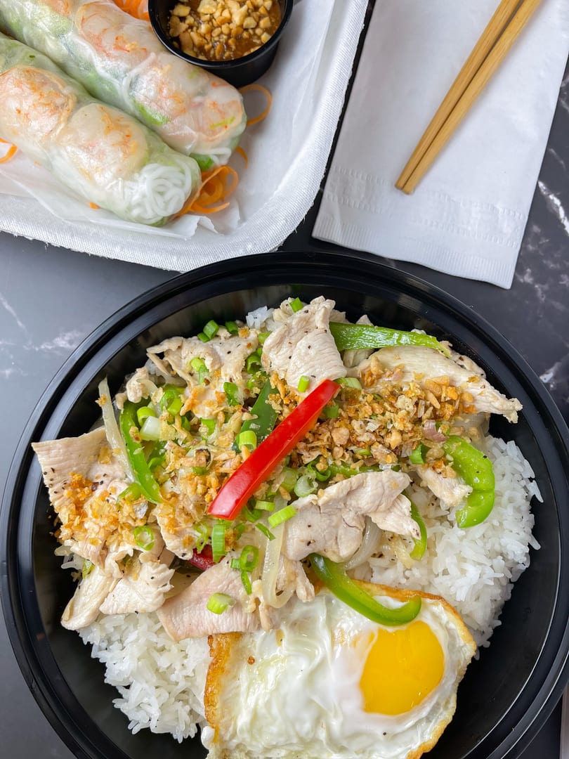 Chicken and rice bowl with a fried egg, spring rolls, and peanut sauce on a dark surface.