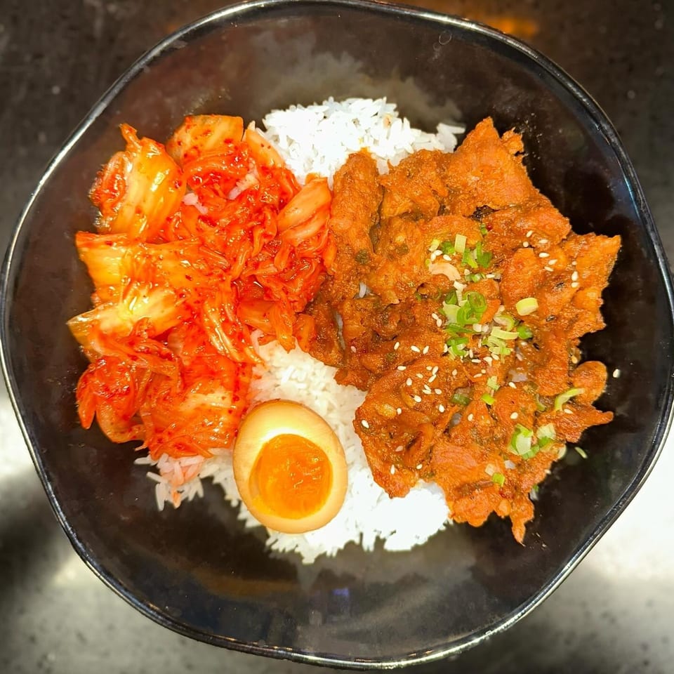 A black bowl filled with white rice, spicy red kimchi, marinated pork, and a soft-boiled egg.