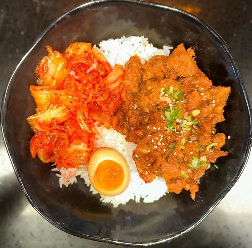 A black bowl filled with white rice, spicy red kimchi, marinated pork, and a soft-boiled egg.