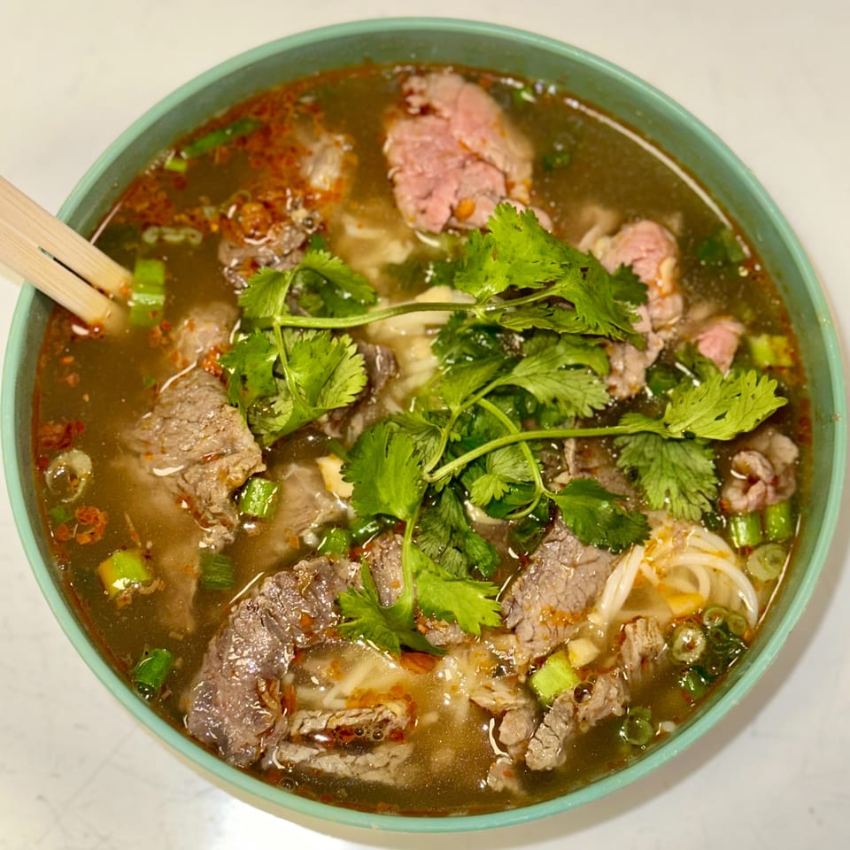 Close-up of a bowl of pho with beef, noodles, cilantro, and green onions, with chopsticks.
