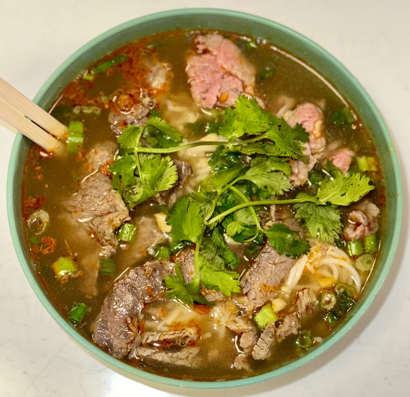 Close-up of a bowl of pho with beef, noodles, cilantro, and green onions, with chopsticks.